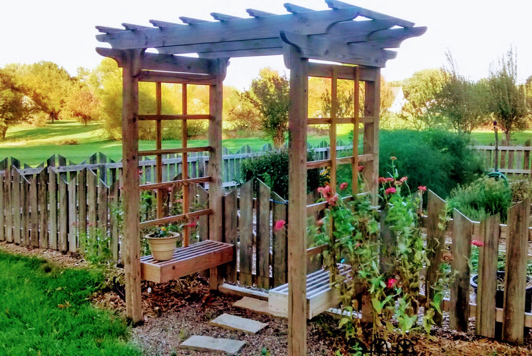 Wooden garden arbor with a trellis, built-in bench, and pink flowers beside a mowed lawn.