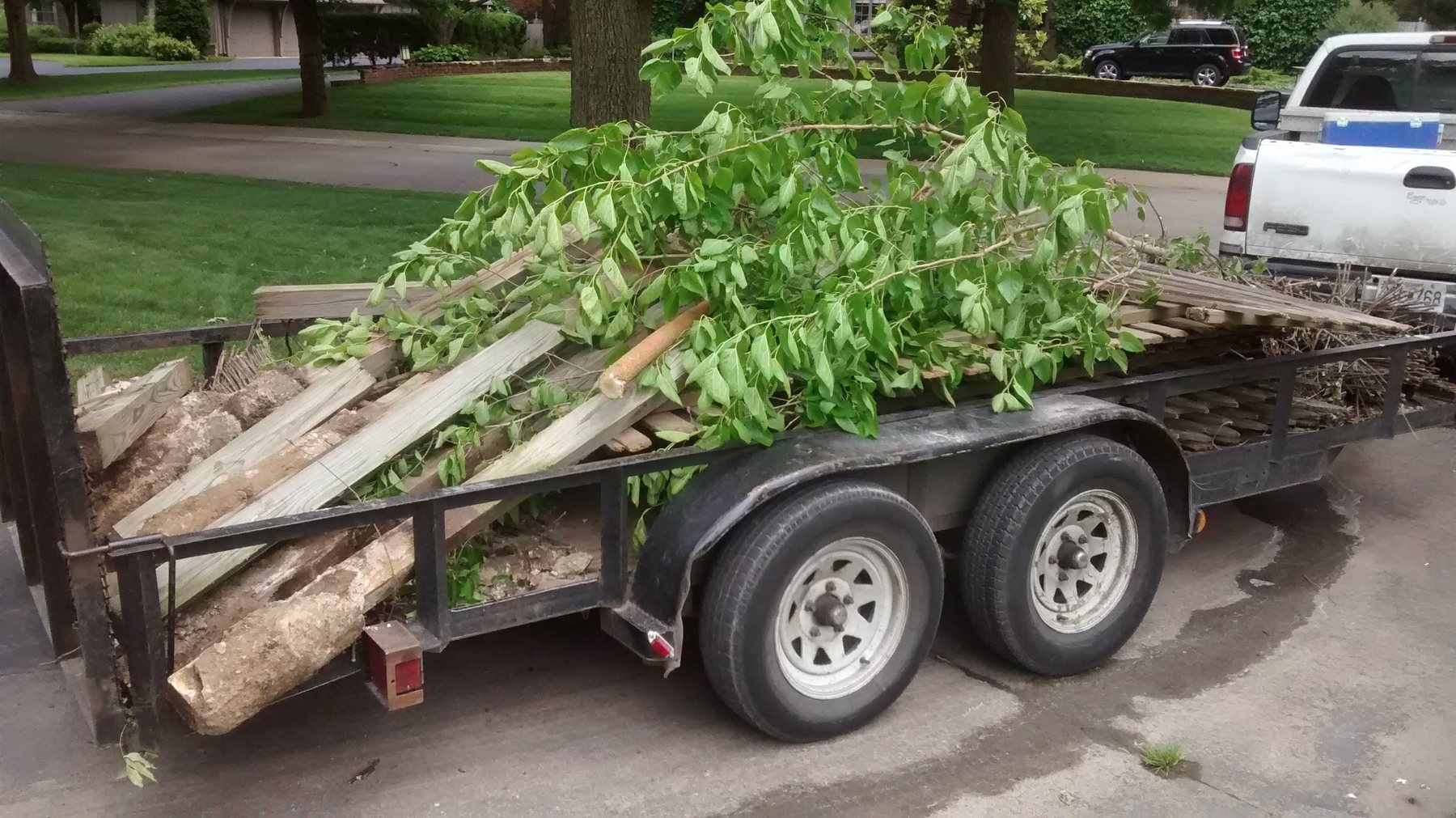 Utility trailer loaded with branches, fence boards, and stone, hitched to a pickup.